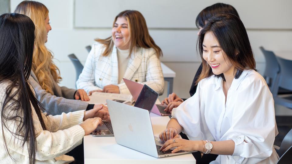 A female in professional clothing is smiling at a laptop while other around her talk